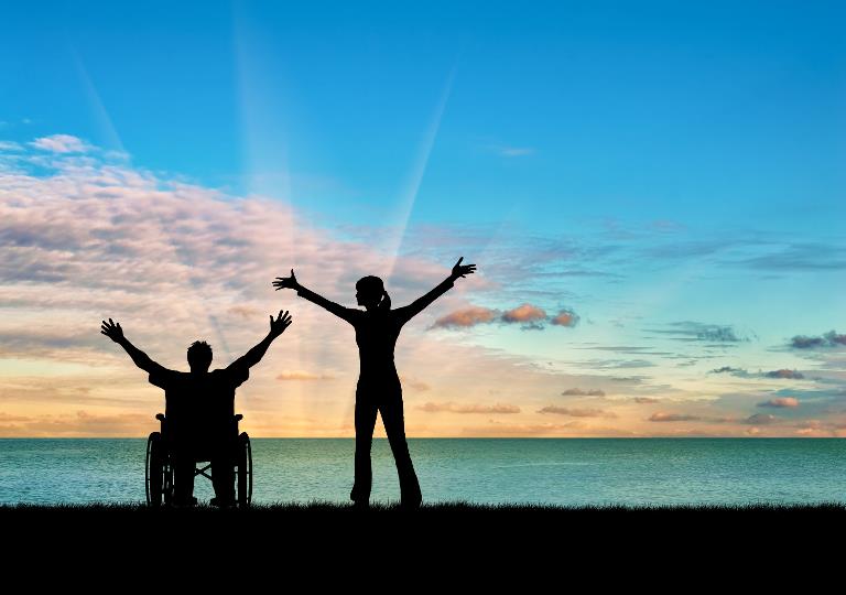 En kørestolsbruger og en stående person med armene i vejret i silhouet foran en solnedgang ved havet 